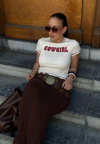 Hvid t-shirt med rød "COWGIRL" skrift, brun tekstureret maxiskørt, stor metalbælte og mørke solbriller. Sidder på stentrappen.