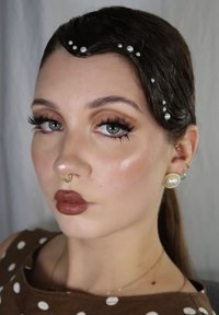 Modella con lunghi capelli adornati da piccole perle, indossa rossetto bronzo, grandi orecchini di perle e un anello al setto d'oro, su uno sfondo grigio.