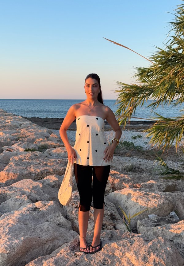 Femme en haut blanc à pois et pantalon noir se tient sur une rive rocheuse au coucher du soleil, tenant un sac blanc avec la mer et un palmier en arrière-plan.