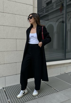 Abrigo largo negro sobre una camiseta sin mangas blanca con un logo, combinado con pantalones anchos negros y zapatillas blancas con detalles negros.