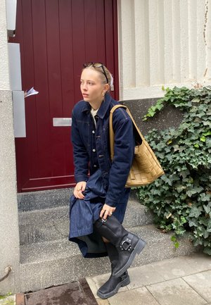 Denimkleid mit langen Ärmeln und Taschen, kombiniert mit schwarzen kniehohen Stiefeln und einer gewebten, tanfarbenen Handtasche. Der Hintergrund zeigt Efeu und Treppen.