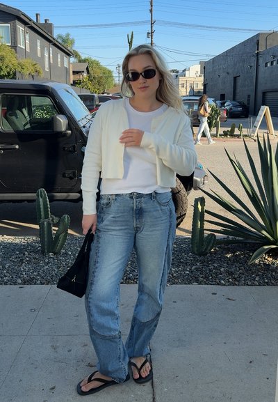 Joven mujer con gafas de sol, cárdigan blanco, vaqueros y chanclas, de pie en la acera cerca de un SUV negro estacionado y plantas desérticas.