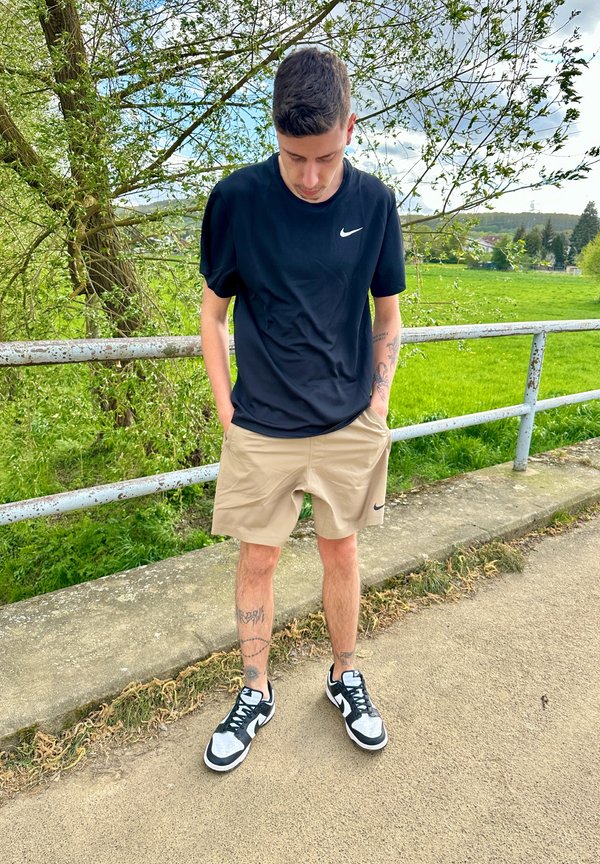 Camiseta negra de Nike de manga corta, pantalones cortos beige de Nike y zapatillas negro y blanco. De pie en un camino con vegetación y una valla de fondo.