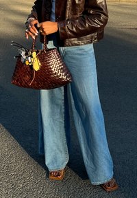 Brunnet vevd håndveske med to håndtak; blå vide denimjeans; brune sandaler; fingre prydet med ringer; teksturert skinnjakke.