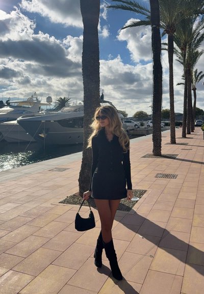 Vestido largo negro de manga larga con botones, botas negras hasta la rodilla y un pequeño bolso negro. Al fondo se ven palmeras y yates.