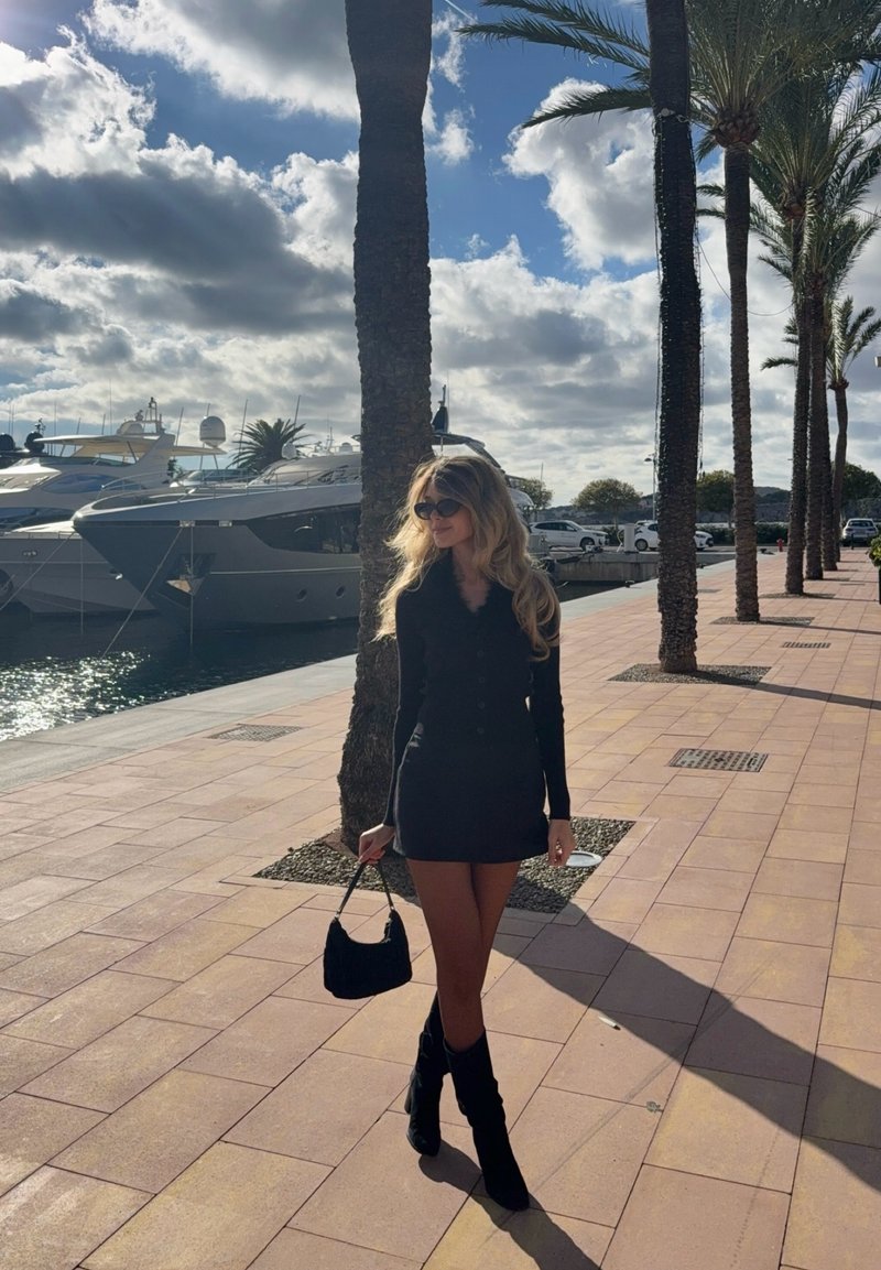Robe noire à manches longues avec boutons, bottes noires montantes jusqu'au genou, et un petit sac à main noir. Des palmiers et des yachts sont visibles en arrière-plan.