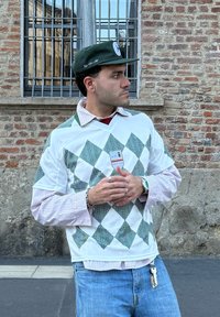 Zeleno-bela pulover z argyle vzorcem, носен preko dolgih črtastih rokavov. Temno zelena kapa z logotipom. Ključevec viden na kavbojkah.