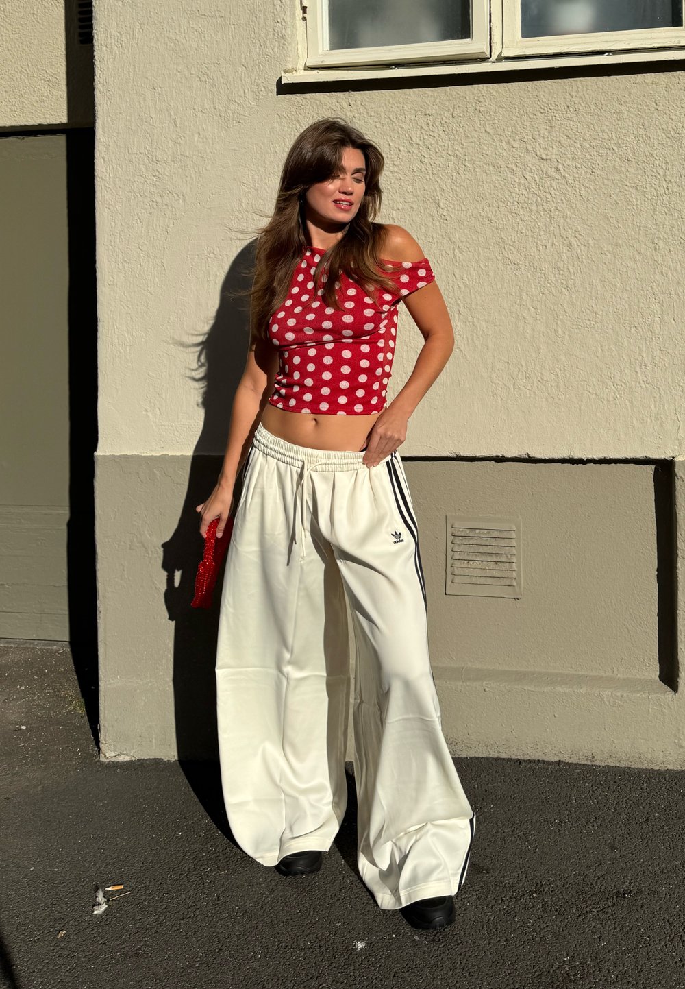 Mulher com um top off-shoulder de bolinhas vermelhas e calças largas brancas da Adidas, de pé contra uma parede bege ao sol, segurando uma pequena bolsa vermelha.