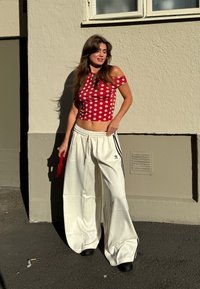 Mulher com um top off-shoulder de bolinhas vermelhas e calças largas brancas da Adidas, de pé contra uma parede bege ao sol, segurando uma pequena bolsa vermelha.