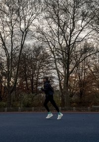 Fekete sportdzseki, fekete nadrág, fehér futócipők t�ürkiz részletekkel. Kék futópályán fut, háttérben kopár fák.