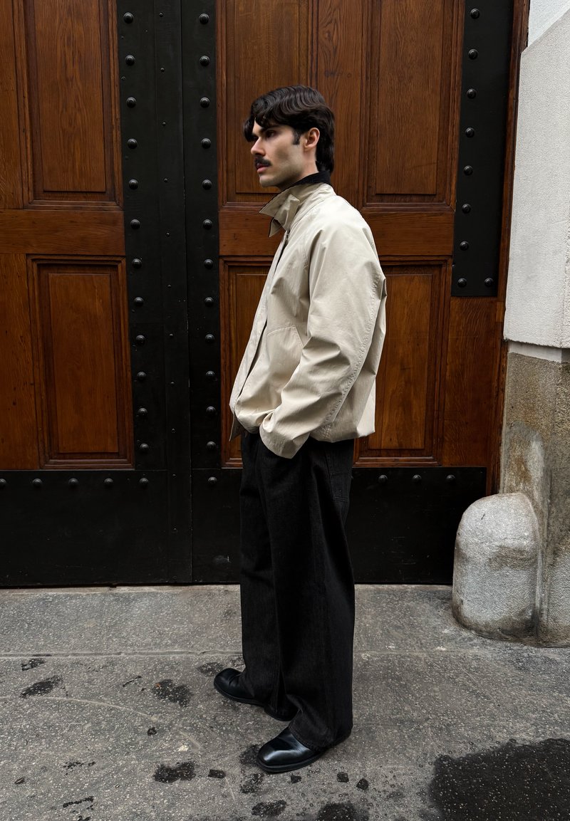 Veste aviateur beige à col montant, associée à un pantalon noir large et des chaussures noires, devant une porte en bois.