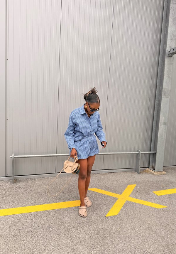 Femme en robe à carreaux bleue et chaussures beige debout sur un asphalt avec des marques jaunes en X, tenant un petit sac à main beige contre un mur en métal.