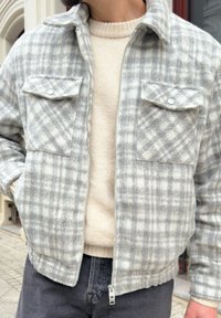 Giacca a zip grigia e bianca a quadri con due tasche sul petto, realizzata in un tessuto morbido e strutturato, abbinata a un maglione color crema sottostante.