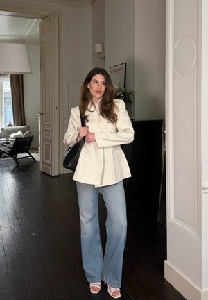 Mujer con cabello largo y castaño, vistiendo un blazer crema, jeans azul claro y sandalias blancas, sosteniendo un bolso negro de hombro en una habitación moderna.