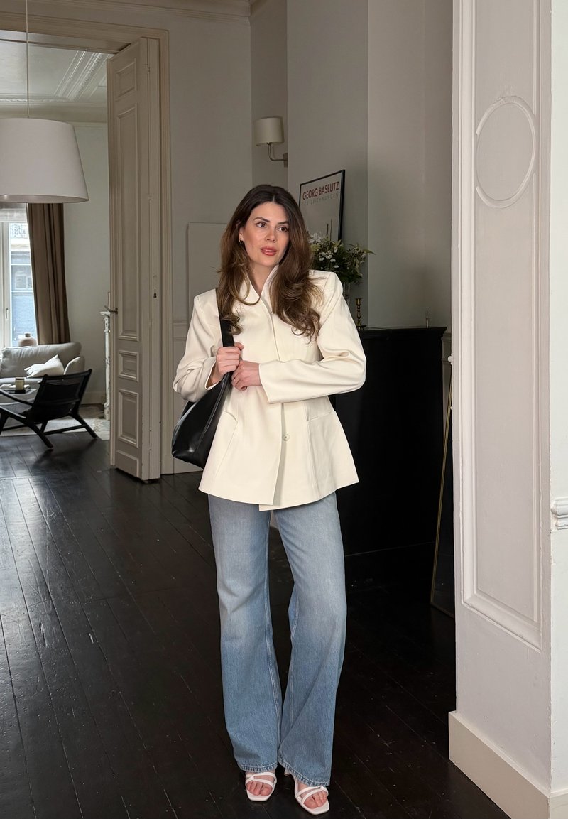 Femme aux longs cheveux bruns portant un blazer crème, un jean bleu clair et des sandales blanches, tenant un sac à bandoulière noir dans une pièce moderne.