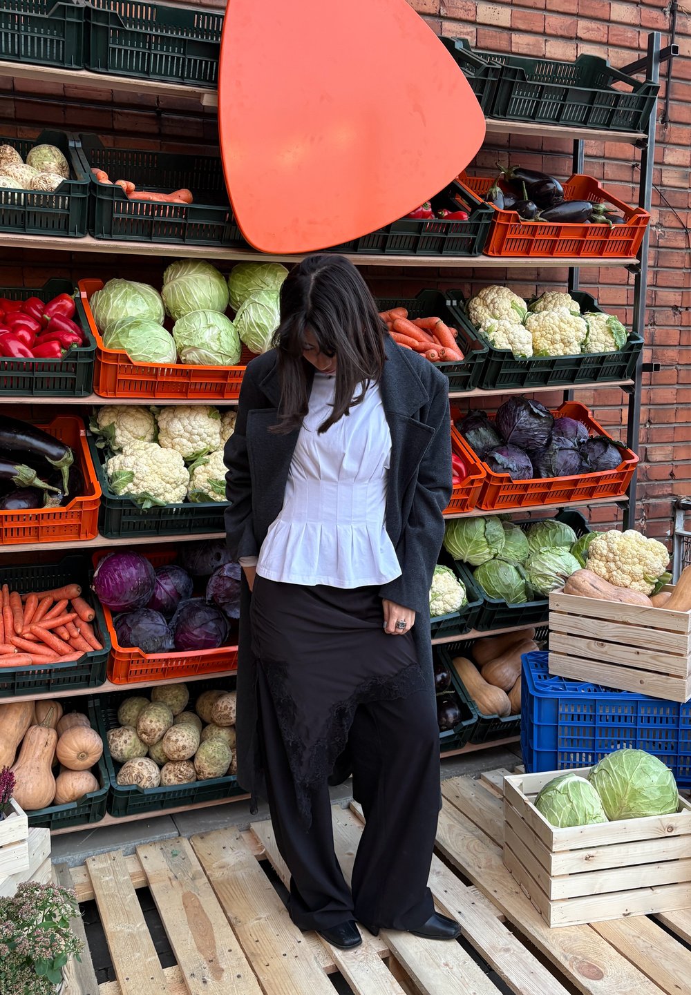 Pantalon noir à jambes larges et haut péplum blanc, accessoirisés d'une bague, posés devant un étal de légumes. Paniers de produits orange et vert visibles.