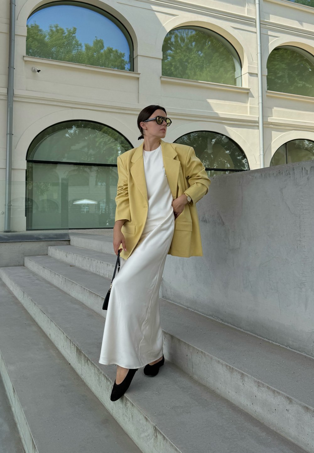 Dzeltena pārmērīga žakete, balta satīna kleita, melni norādīti zābaki, turēdama melnu rokassomu, uz pelēkām betona pakāpēm, mūsdienīgas arhitektūras fons.