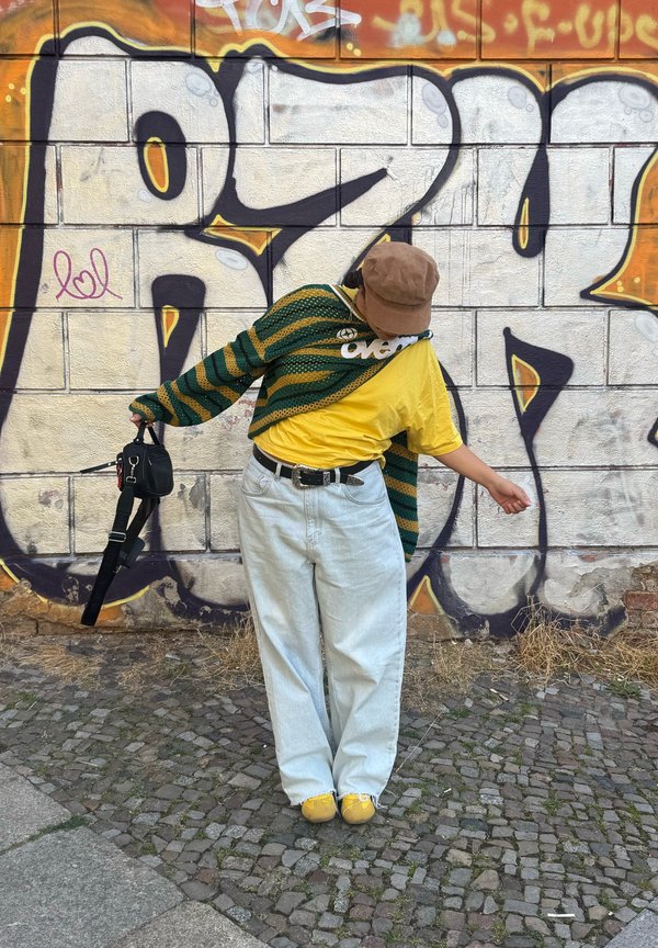 Persoon draagt een bruine pet, gele blouse, gestreepte groene trui, witte wijde broek en gele schoenen, staat tegen een graffitimuur en houdt een zwarte tas vast.