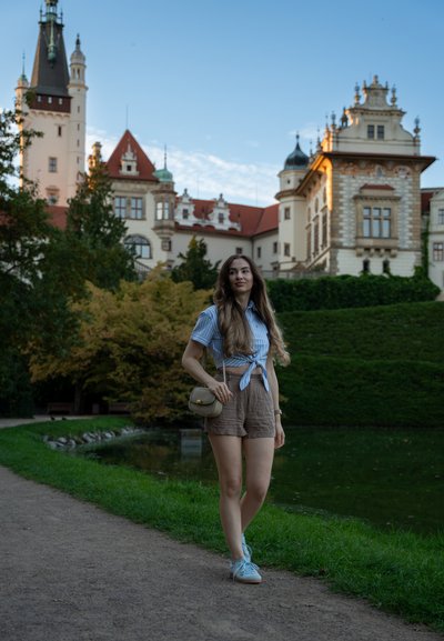Camisa de rayas azul claro de manga corta anudada a la cintura, pantalones cortos marrones, zapatillas azul claro y un pequeño bolso de hombro beige junto a un estanque.