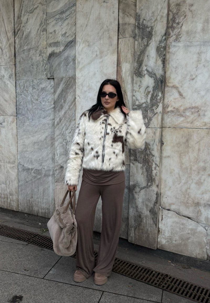 Donna che indossa occhiali da sole, un cappotto di pelliccia sintetica bianca e marrone a macchie, pantaloni larghi marroni, mentre tiene una grande borsa beige, in piedi accanto a una parete di marmo.