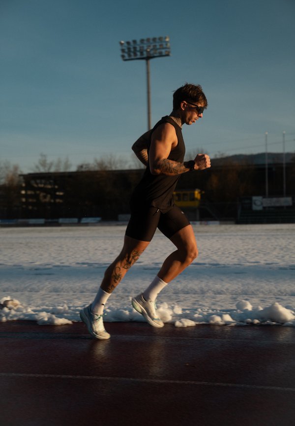 Moški s tetovažami in sončnimi očali, ki teče po stezi, oblečen v črno športno obleko in bele nogavice, s snegom na polju ob stezi.