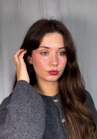 Grå texturerad tröja med knappdetaljer, långt vågigt brunt hår och en septumring. Fräknig hy och naturlig smink förstärker utseendet.