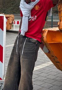 Raudona Manchester United marškinėlių su logotipu, pilkas megztinis aptrauktas aplink kaklą ir laisvos tamsiai mėlynos džinsinės kelnės su baltomis raištelėmis. Betono fonas.
