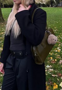 Blondínka v čiernom oblečení si upravuje golier kabáta, nesie olivovozelenú tašku s plyšovým medveďom na kľúčenke, stojí na tráve s popadaným lístím.