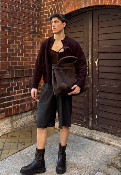 Bolso tote de ante marrón oscuro con forma estructurada y asas curvas, combinado con un cárdigan oscuro de punto y pantalones cortos grises.