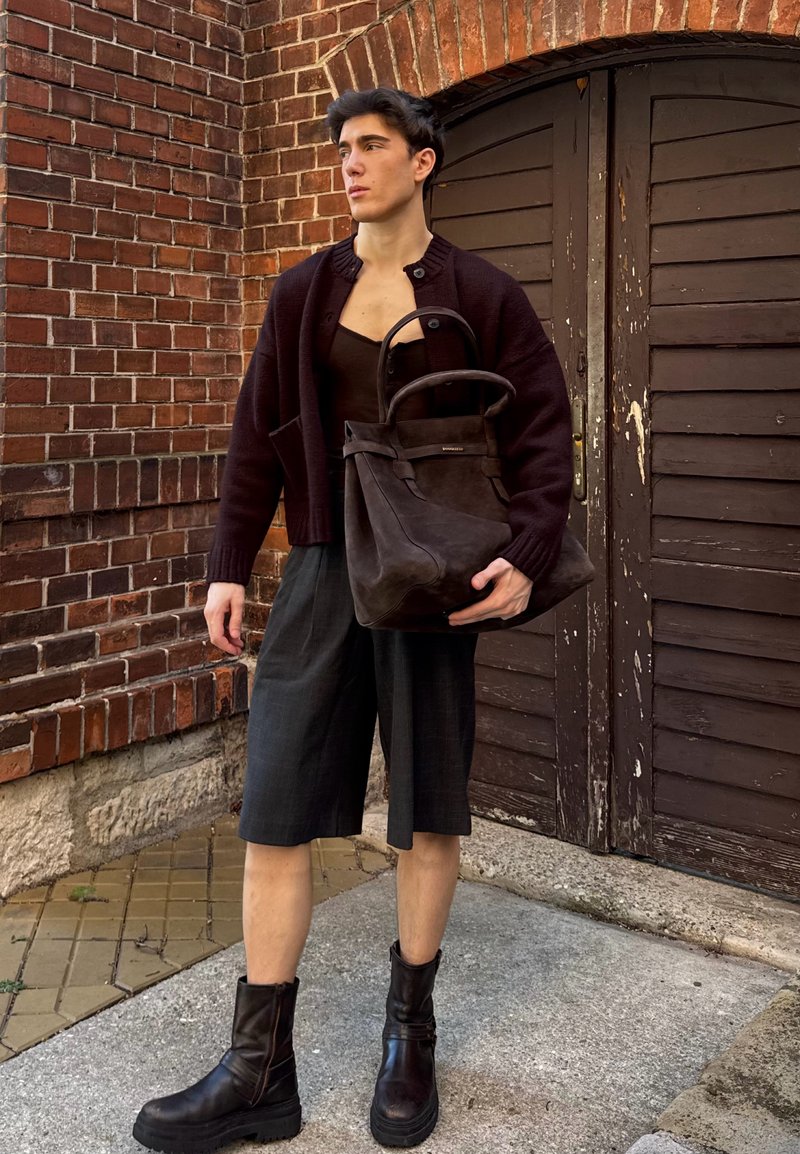 Dark brown suede tote bag with a structured shape and curved handles, paired with a knitted dark cardigan and grey shorts.