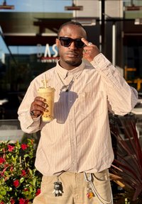 Vīrietis ar saulesbrillēm un bēša krāsas svītrainu kreklu tur rokā burbuļtēju ar salmiņu, stāv ārā pie augiem un ēkas.