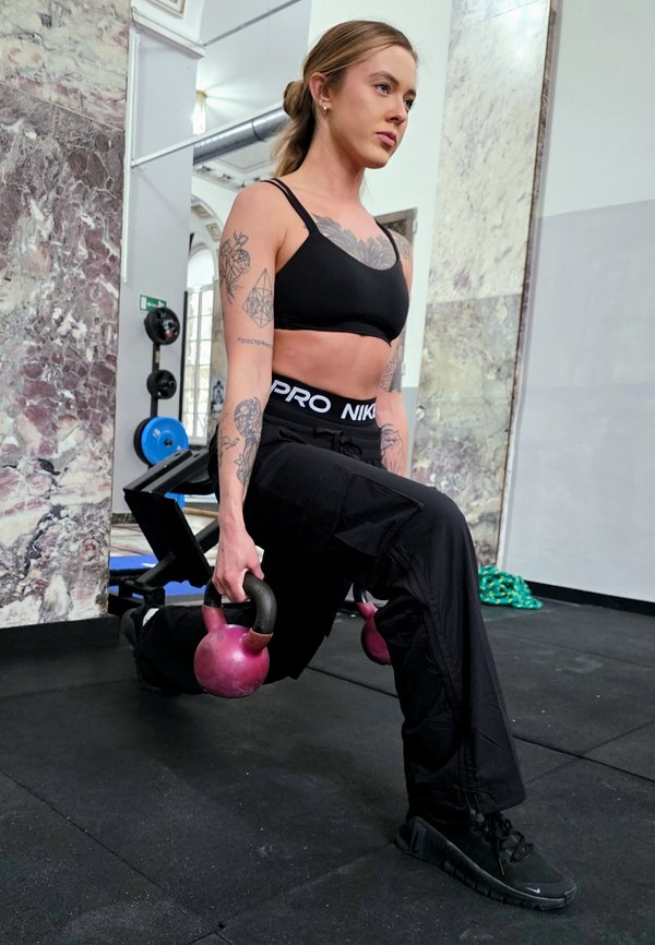 Veltrent kvinne med tatoveringer utfører vektet utfall mens hun holder rosa kettlebells i treningsstudio med marmormurer og treningsutstyr.