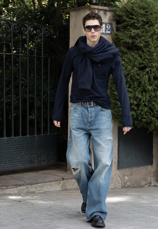 Joven con vaqueros extragrandes, zapatos negros, camisa azul marino con suéter sobre los hombros y gafas de sol caminando por una acera al aire libre.