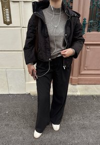 Musta tikattu takki harmaan korkeakauluksisen neuleen päällä, yhdistettynä löysiin mustiin housuihin ja valkoisiin jalkineisiin. Kuulokkeet ja älypuhelin näkyvissä.