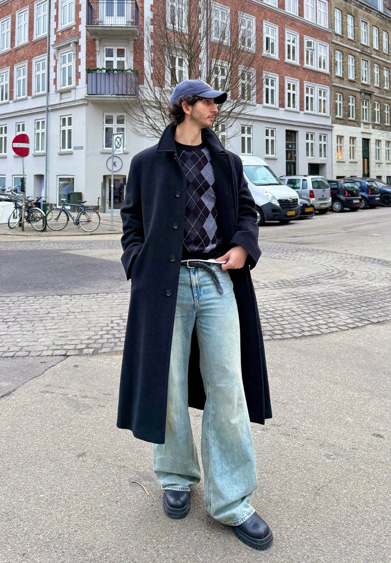 Jeune homme portant un long manteau noir, un pull à losanges, un jean clair ample, des chaussures noires et une casquette sombre, debout dans une rue de la ville avec des bâtiments et des voitures garées.
