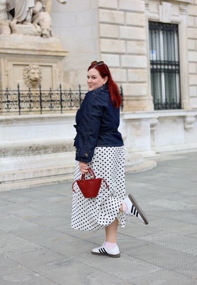 Mujer con cabello rojo con chaqueta vaquera y falda de lunares, sosteniendo un bolso rojo, levanta un pie en una calle de la ciudad con edificios de piedra.