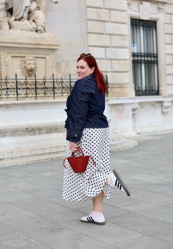 Frau mit roten Haaren in einer Jeansjacke und gepunktetem Rock, hält eine rote Handtasche und hebt einen Fuß auf einer Stadtstraße mit steinernen Gebäuden.
