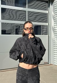 Casaco preto com fecho éclair, textura floral e mangas bufantes, combinado com calças de cintura alta a condizer. Design simples e ajuste descontraído.