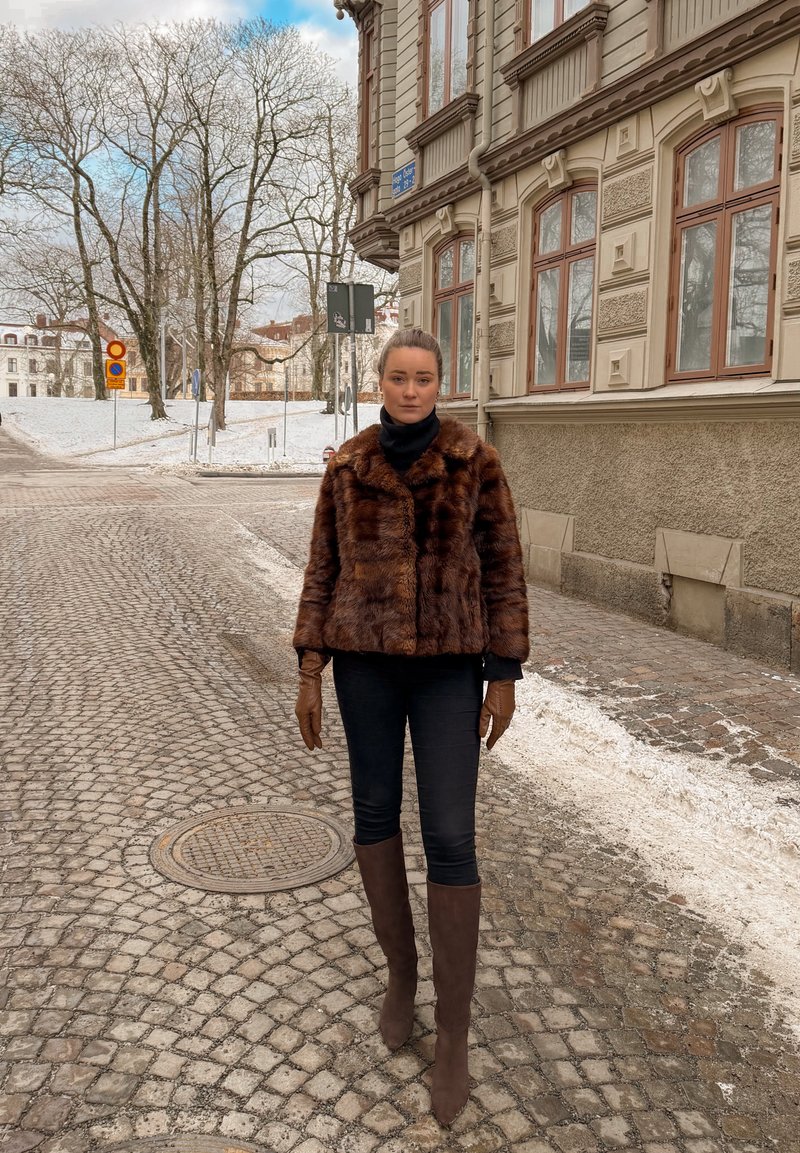 Femme en veste de fourrure marron, gants et bottes marchant sur une rue pavée à côté d'un bâtiment historique par une froide journée d'hiver.