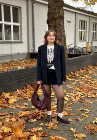 Blazer nero a righe, t-shirt bianca grafica, gonna nera, collant trasparenti, borsa color bordeaux e mocassini con calzini beige tra le foglie autunnali.