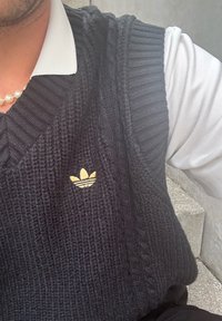 Sweater sem mangas de malha preta com textura canelada, apresentando o logótipo dourado da Adidas. Usado por cima de uma camisa branca com colarinho.