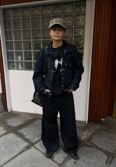 Persona con gorra caqui, chaqueta de mezclilla oscura, camiseta negra con gráfico, pantalones anchos negros y zapatos negros, de pie en una acera de baldosas cerca de una pared de bloques de vidrio.