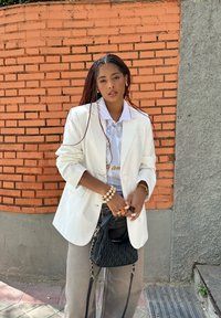 White oversized blazer, light blue shirt featuring gold text, grey trousers, and a black textured handbag; accessorised with multiple rings and a bracelet.