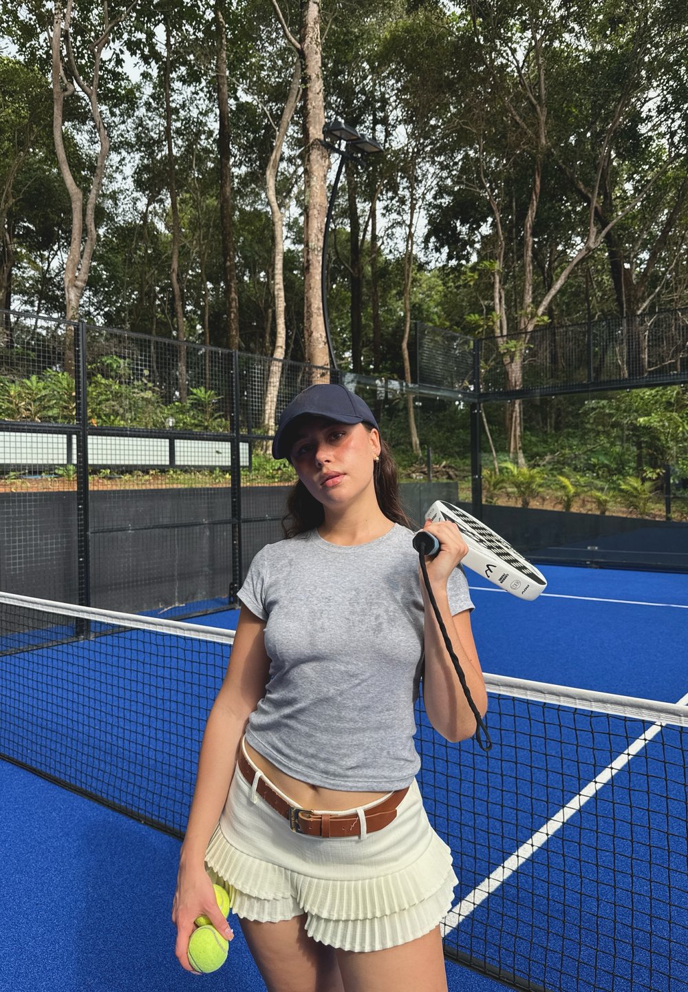 Joueur de tennis tenant une raquette blanche et une balle de tennis jaune, portant un maillot gris et un short plissé crème, sur un court bleu avec des arbres.