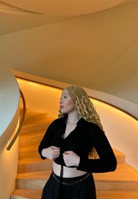 Svart långärmad crop top med volangdetalj vid halsen, kombinerad med en svart kjol med dekorativ midja. Inomhustrappa i bakgrunden.