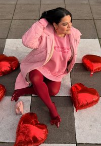 Personne en manteau rose et pull avec le texte « Romanticize Your Life », assise sur un pavement à motifs entourée de ballons rouges en forme de cœur.