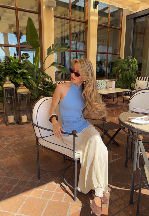 Mujer con top azul sin mangas y falda blanca sentada en una silla blanca acolchada en una cafetería al aire libre con plantas y ventanas de fondo.
