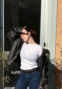 Czarna skórzana kurtka na białym prążkowanym topie, niebieskie dżinsy i ozdobny pasek. Modelka pozuje przy wejściu do budynku. Tło oświetlone słońcem.