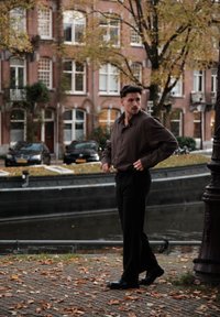 Braunes Langarmhemd, schwarze Hose und schwarze Schuhe. Der Hintergrund zeigt einen Kanal, Backsteingebäude und Herbstblätter auf dem Boden.