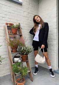 Schwarzer Blazer über einem weißen T-Shirt, kombiniert mit schwarzen Radshorts. Beiger Handtasche, gemusterten Sneakern und einem hölzernen Pflanzenständer mit Blumen.